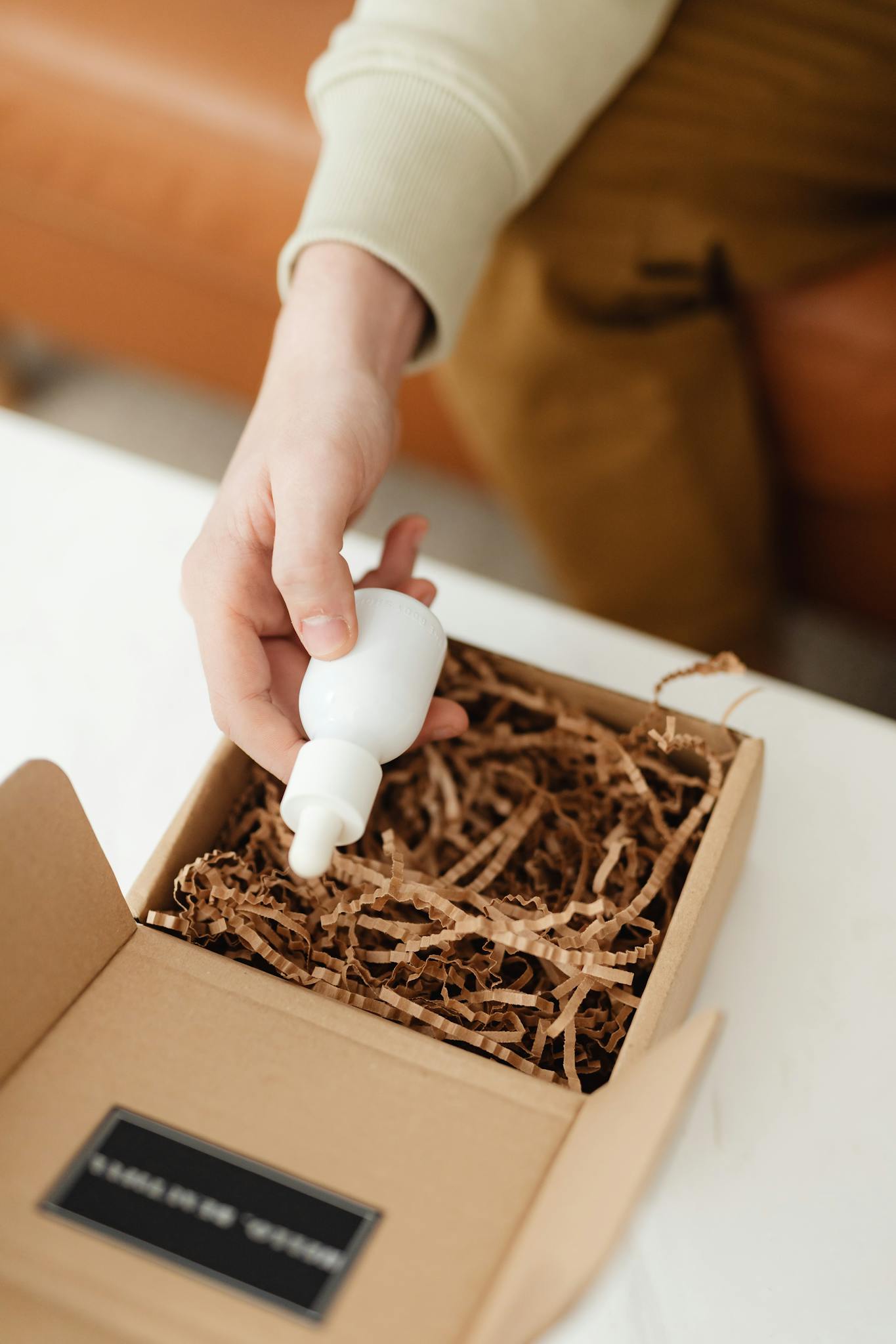 A person holding a dropper bottle over an open cardboard box with packing material inside.