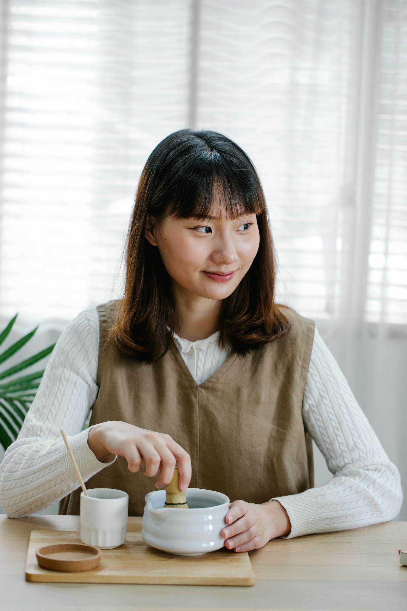 Asian woman seated indoors, smiling while holding a ceramic bowl and cup with natural light.